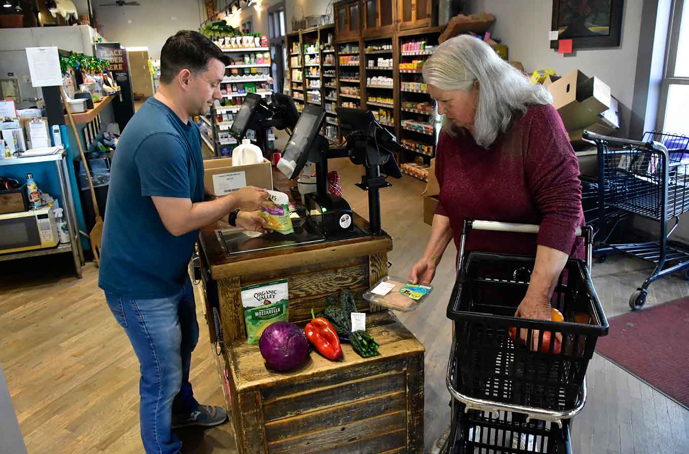 A cashier scans groceries for an older woman with a shopping cart at a small natural food store checkout counter.