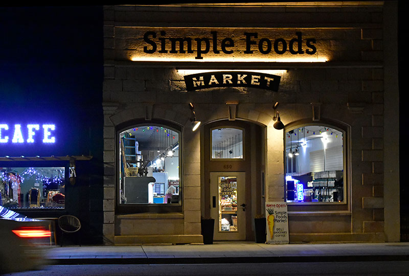 The exterior of Simple Foods Market in Del Norte, Colorado, photographed at night with the storefront lit up and an open sign visible in the window.