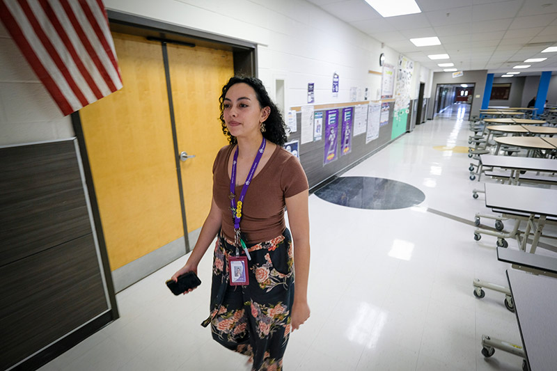 A school staff member wearing a lanyard walks through a hallway lined with lunch tables.
