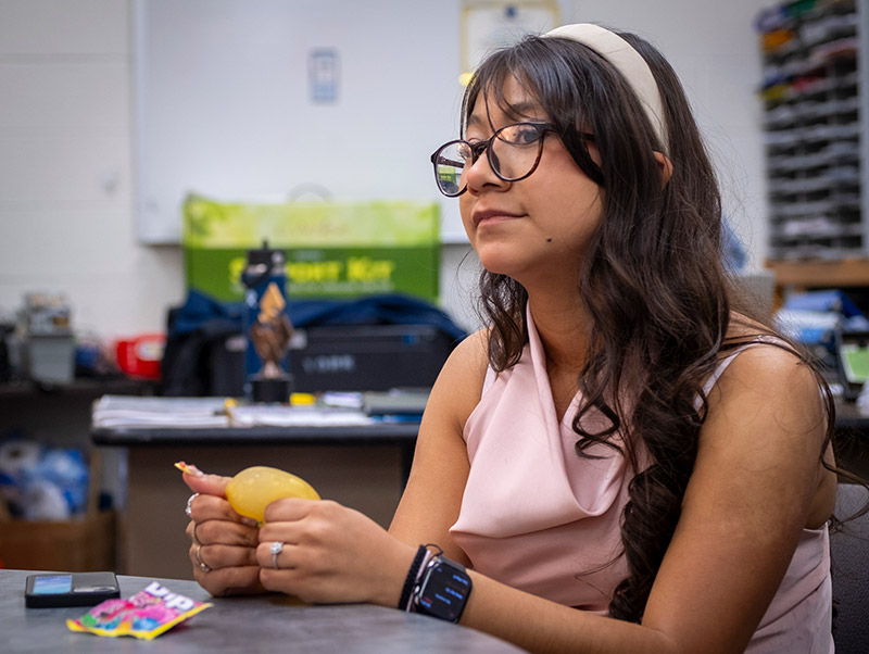 A young woman sits at a table holding a yellow stress ball, looking off to the side in thought.
