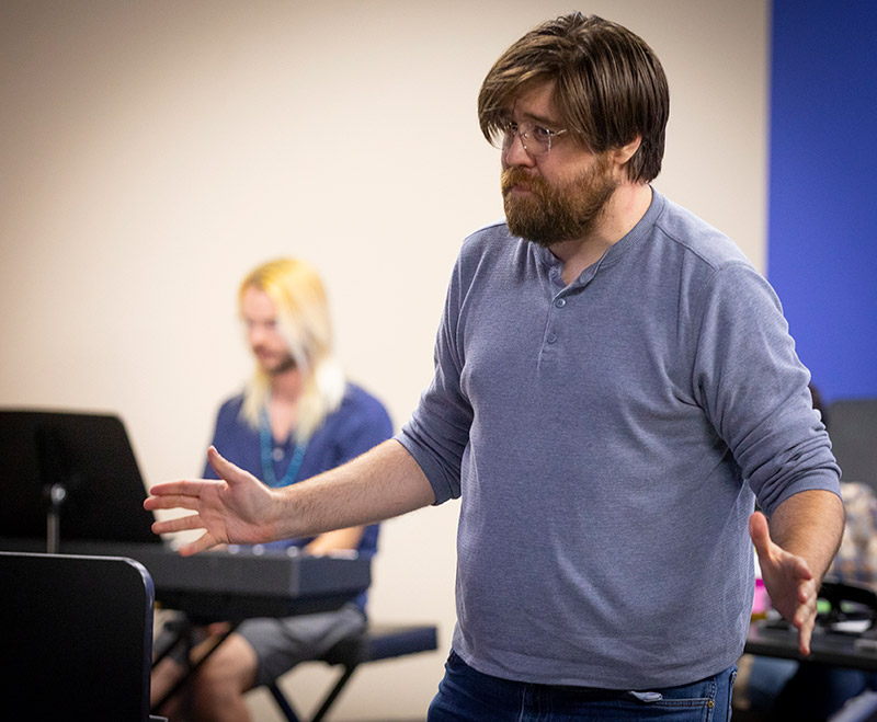 A bearded man in a gray henley gestures with both hands while speaking, with a musician playing keyboard in the background.