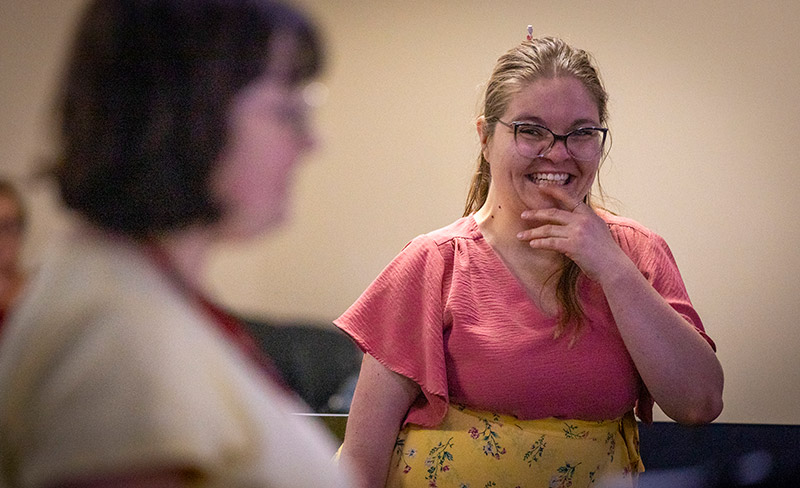 A woman in a pink top and yellow floral skirt laughs mid-conversation, hand near her chin, with another woman blurred in the foreground.