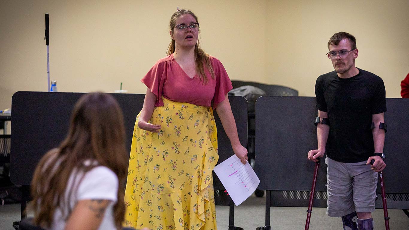 A woman in a pink top and yellow skirt speaks while holding sheet music, standing near a man using forearm crutches, with another person seated in the foreground.